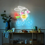 Bunny on Bike Easter LED neon sign glowing on wall with playful bunny riding a bike design for home decor, nursery, party, or shop display by Neon.Shop.