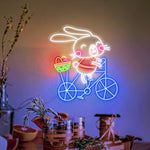 Bunny on Bike Easter LED neon sign glowing on wall with playful bunny riding a bike design for home decor, nursery, party, or shop display by Neon.Shop.