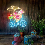 Bunny on Bike Easter LED neon sign glowing on wall with playful bunny riding a bike design for home decor, nursery, party, or shop display by Neon.Shop.